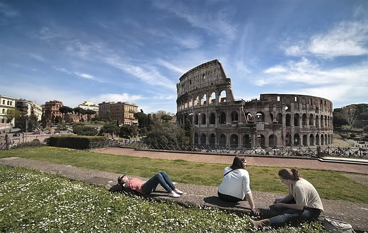 The Colosseum under a blue sky, with visitors sitting on the grass in front of it