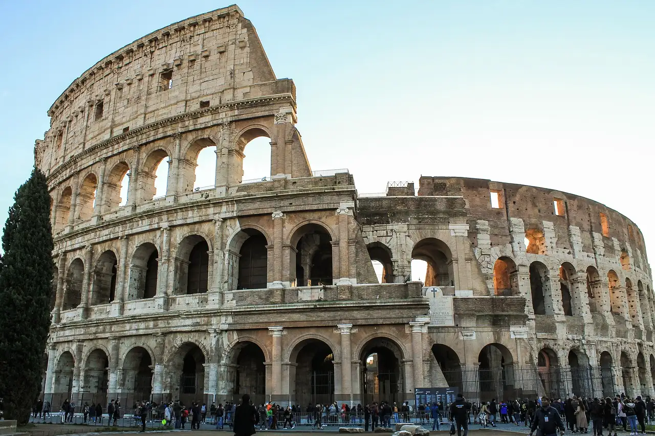 The outer wall of the Colosseum in Rome on a clear day, with a cypress tree on the left