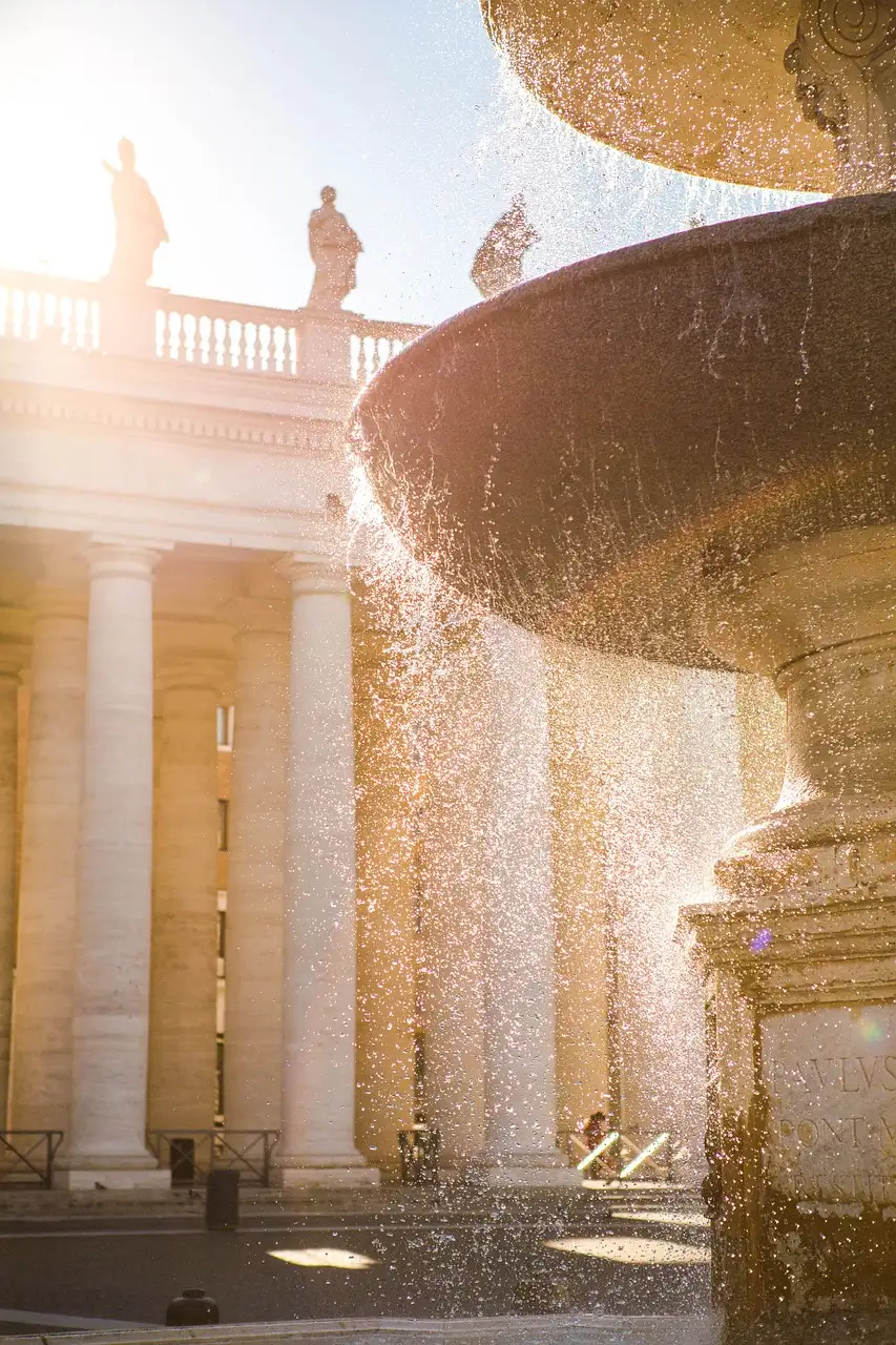 A Roman fountain with water spraying in sunlight beside a stone colonnade