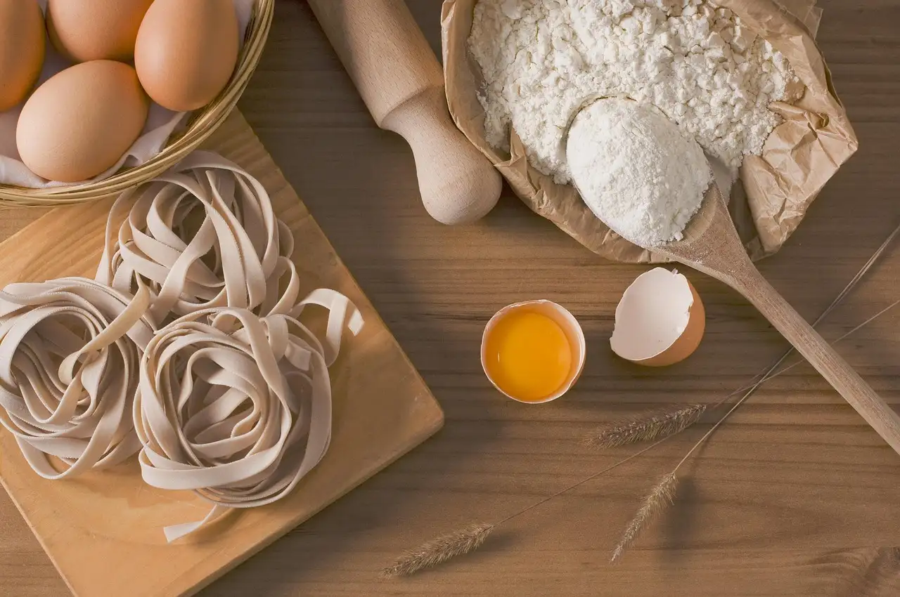 Nests of fresh tagliatelle next to flour, eggs and a rolling pin on a wooden table