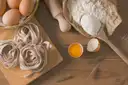 Nests of fresh tagliatelle next to flour, eggs and a rolling pin on a wooden table