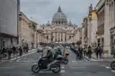 A busy Rome street with scooters and pedestrians, leading to St. Peter's Basilica