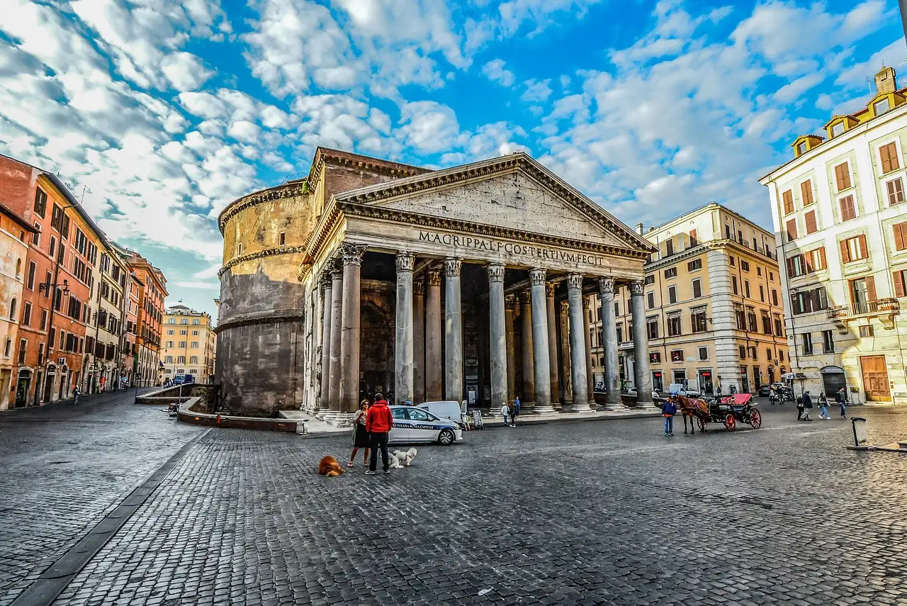 The Pantheon's columns seen from its cobbled square under a bright blue sky with clouds