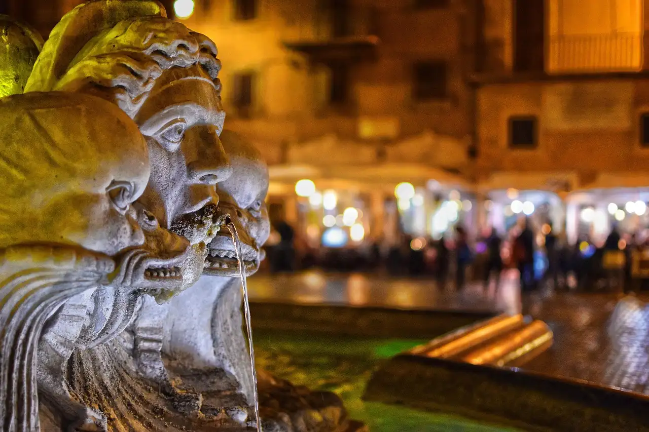 A carved stone face on a fountain in Piazza Navona at night, with lit cafés behind