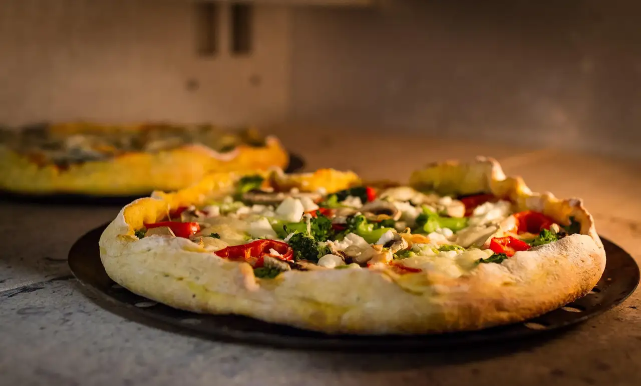 A pizza with vegetables on a stone in front of a pizza oven