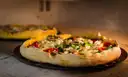 A pizza with vegetables on a stone in front of a pizza oven