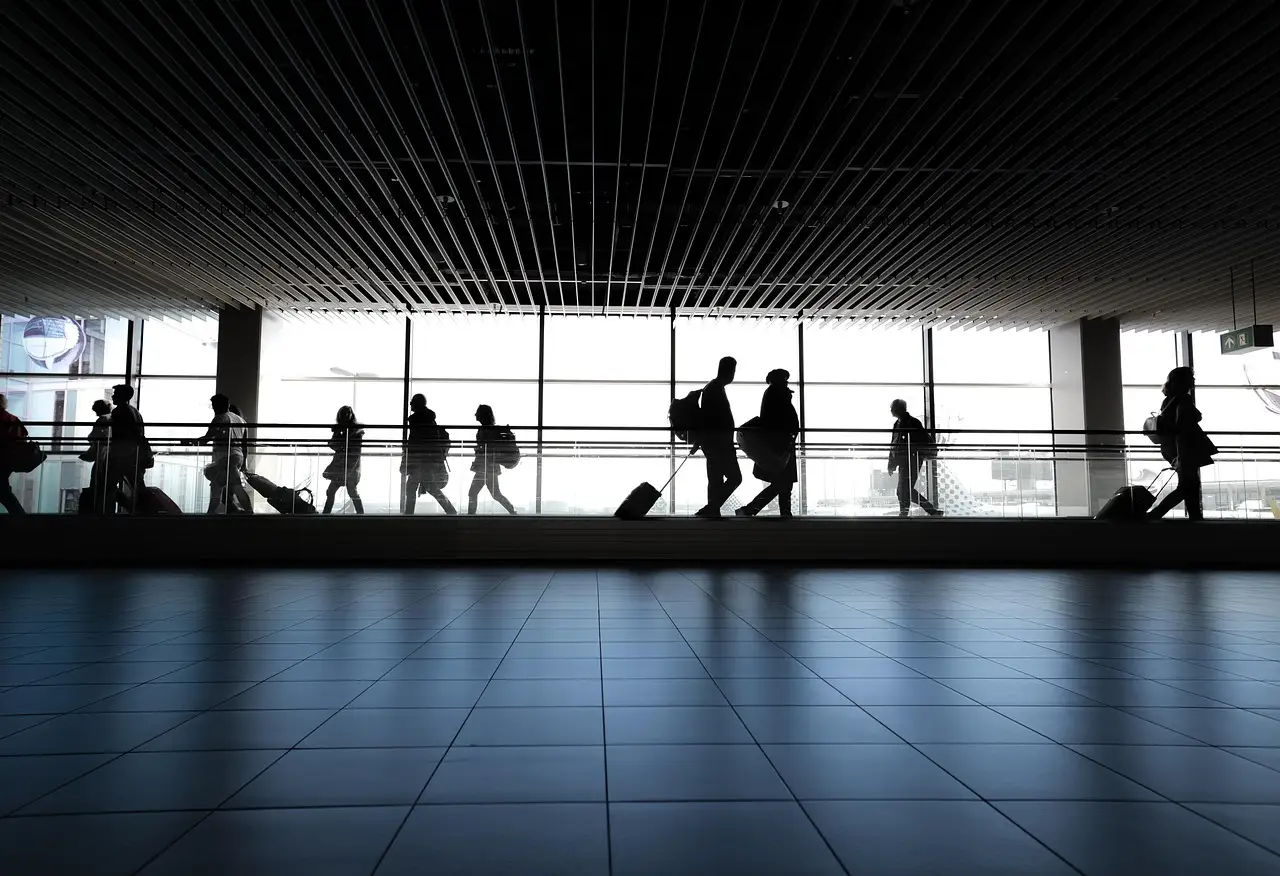 Travellers with wheeled bags walk through a bright airport terminal, seen as silhouettes against tall windows