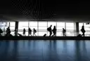 Travellers with wheeled bags walk through a bright airport terminal, seen as silhouettes against tall windows