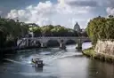 A small boat on the Tiber river with a stone bridge and St. Peter's dome behind