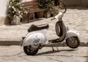 A white vintage scooter parked on a cobbled street by a bench and flower pots
