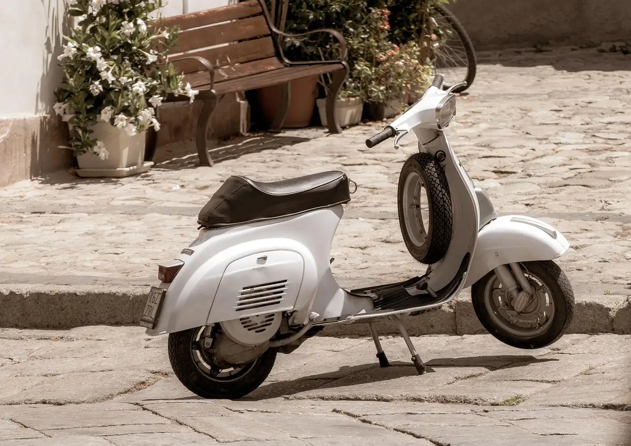 A white vintage scooter parked on a cobbled street by a bench and flower pots